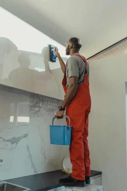 who-we-are A man wearing coveralls is cleaning a large window inside a sleek, modern home interior.